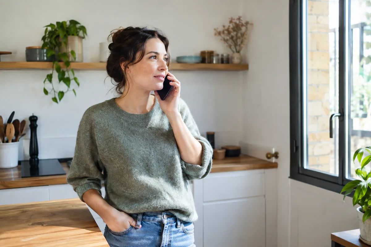 Une adulte fictive téléphone dans sa cuisine calme, sans produit visible.